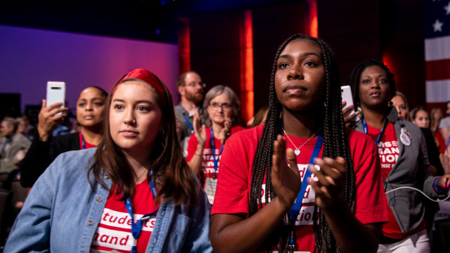 Two Students Demand Action volunteers stand and watch the stage at the Presidential Gun Sense Forum. A crowd of other volunteers in Moms Demand Action shirts stand behind them.