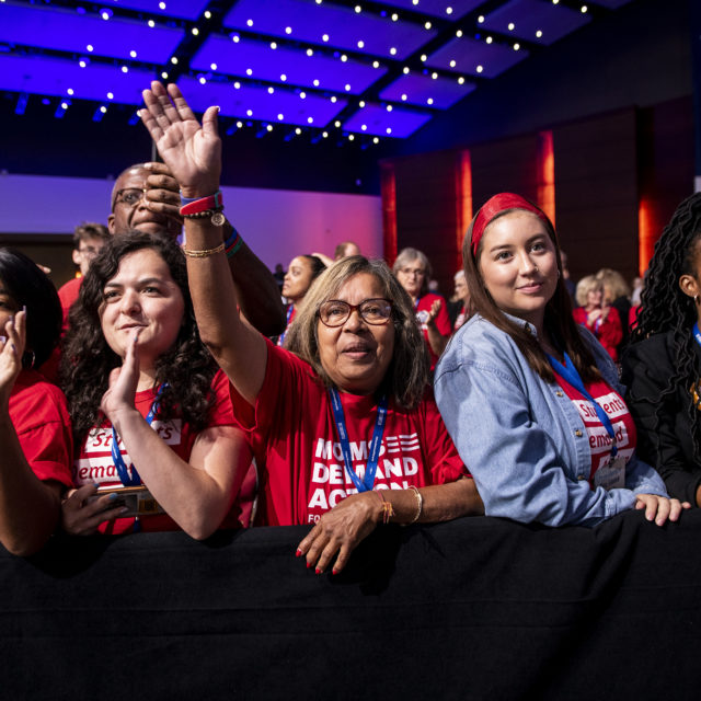 A group of volunteers stand front row at the Presidential Gun Sense forum. Some volunteers raise their hands to wave