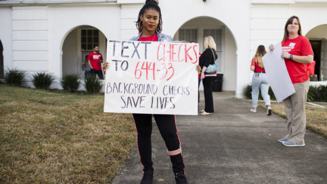 A volunteer stands with a sign saying, 'text checks to 644-33 background checks save lives'