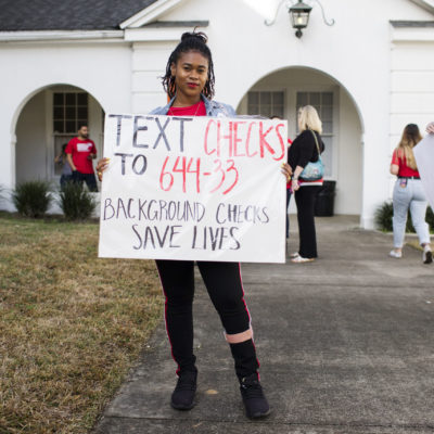 A volunteer stands with a sign saying, 'text checks to 644-33 background checks save lives'