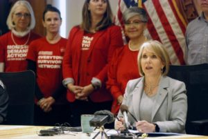 Gov. Lujan Grisham sits and speaks with a microphone in front of her. Behind her, four Moms Demand Action volunteers stand together. 