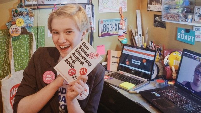 Students Demand Action volunteer Peren Tiemann holds a Gun Sense University sign while smiling and sitting at a computer