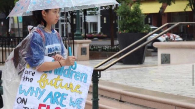 Jeannie stands in the rain with an umbrella while holding a protest sign and wearing a March for Our Lives shirt