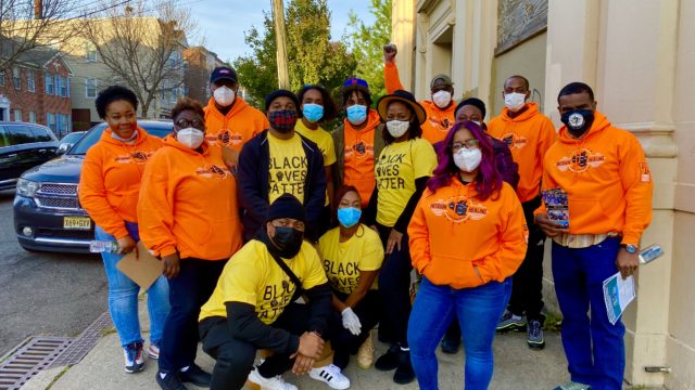 A group of 15 people stand in a group together while wearing Paterson Healing Collective sweatshirts and Black Lives Matter shirts. The group is standing outside while wearing masks during the pandemic.