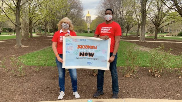 A man and a woman, both wearing Moms Demand Action shirts, hold a Background Checks Now sign on the Road Trip for Background Checks