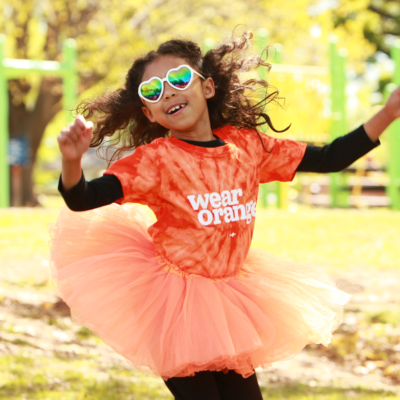 A young girl wearing a Wear Orange shirt and an orange tutu jumps while playing at a public park