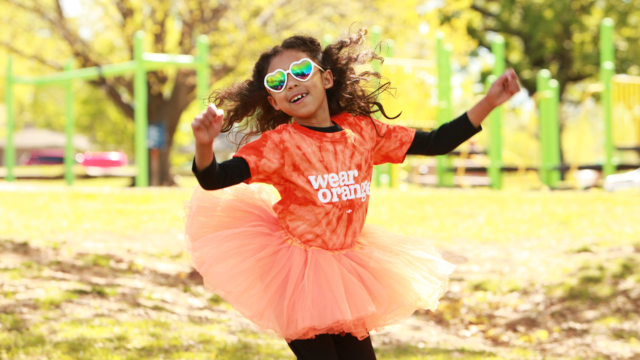 A young girl wearing a Wear Orange shirt and an orange tutu jumps while playing at a public park