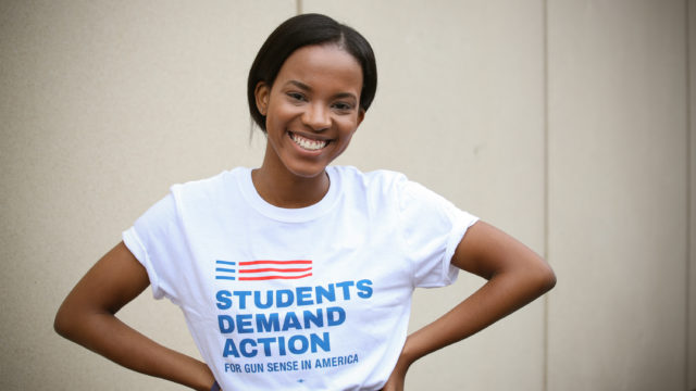 A Students Demand Action volunteer stands and smiles while wearing a Students Demand Action t-shirt