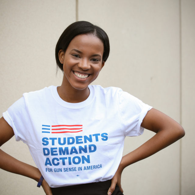 A Students Demand Action volunteer stands and smiles while wearing a Students Demand Action t-shirt