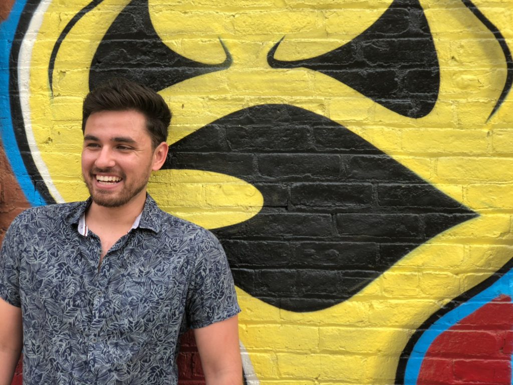 Shane Colombo smiling beside a yellow and black mural