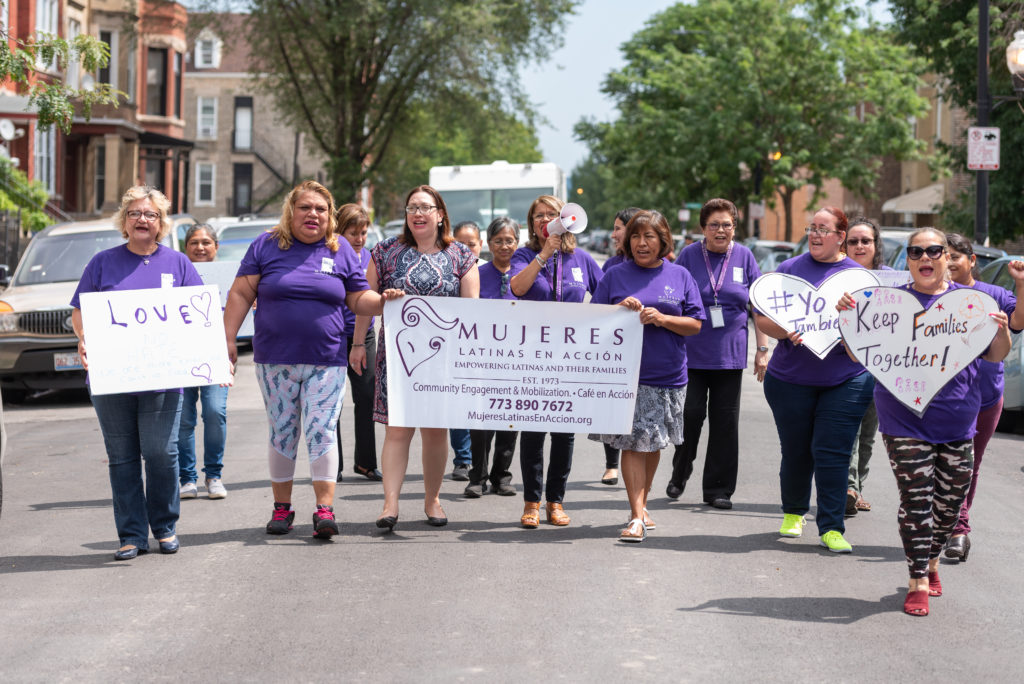 A group of about 15 people march while wearing purple shirts and while holding Mujeres Latinas en Accion signs.