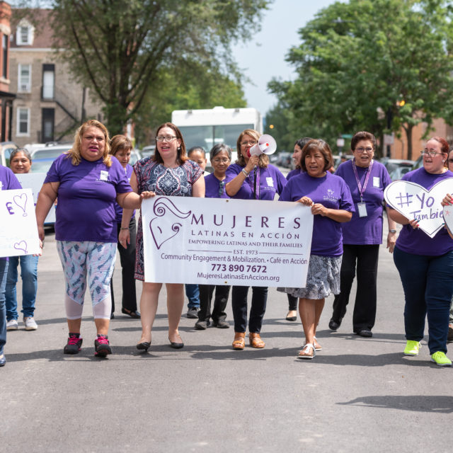 A group of about 15 people march while wearing purple shirts and while holding Mujeres Latinas en Accion signs.