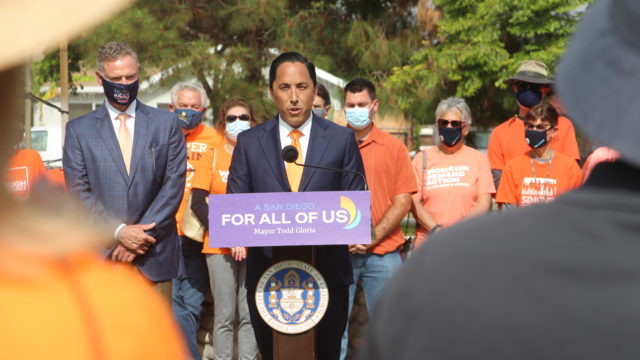 San Diego Mayor Todd Gloria speaks at the podium at an event. Behind him stands a group of people, including Moms Demand Action volunteers