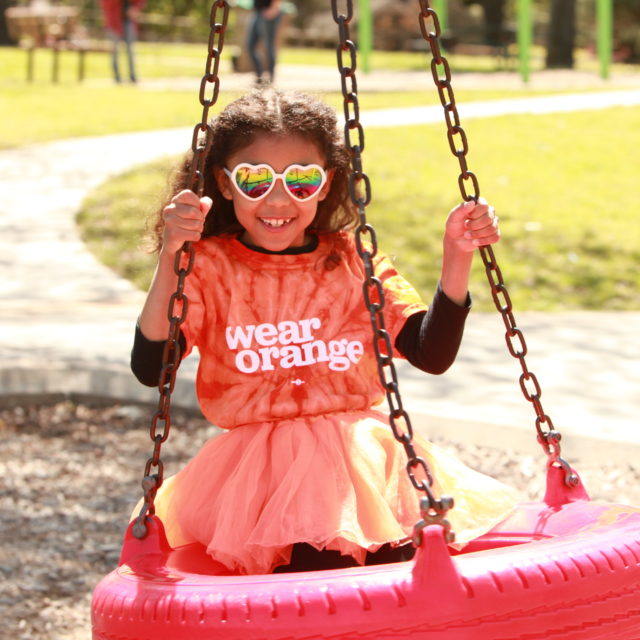 A young girl swings on a tire swing while wearing an orange Wear Orange shirt for National Gun Violence Awareness Day