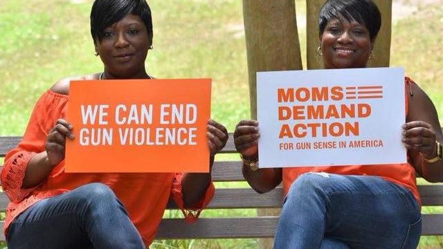 Two women (including Tisa Whack) sit on a park bench holding up signs. One sign is orange and says “We Can End Gun Violence” in white; the other is white and has the Moms Demand Action for Gun Sense in America logo in orange