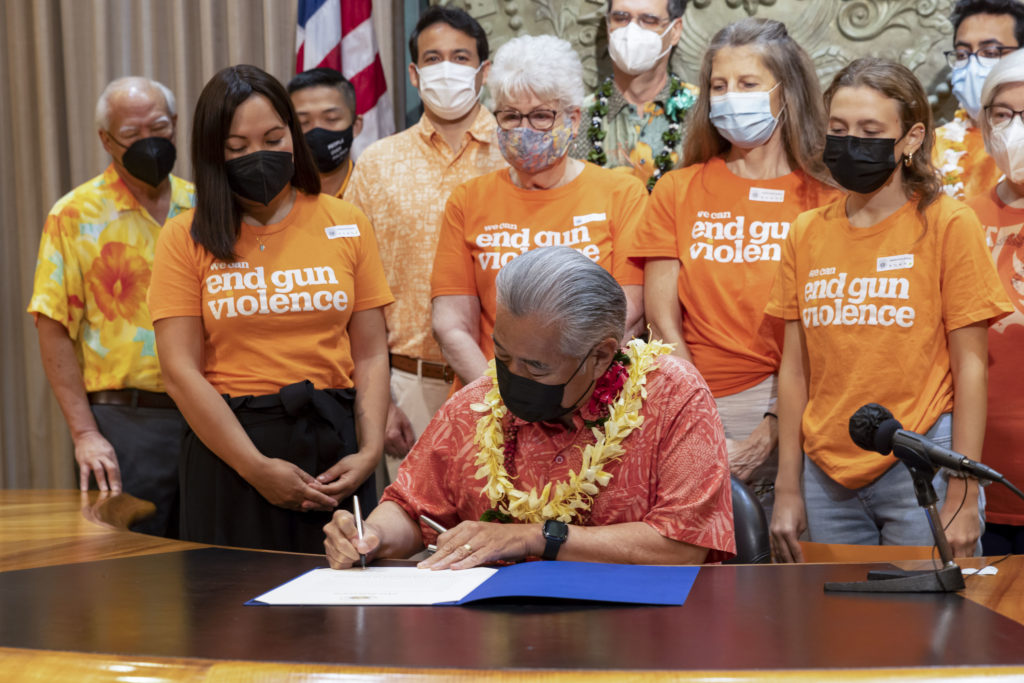 At a table surrounded by people in Wear Orange t-shirts, Hawaii Governor David Ige signs a bill to declare June 3 National Gun Violence Awareness Day