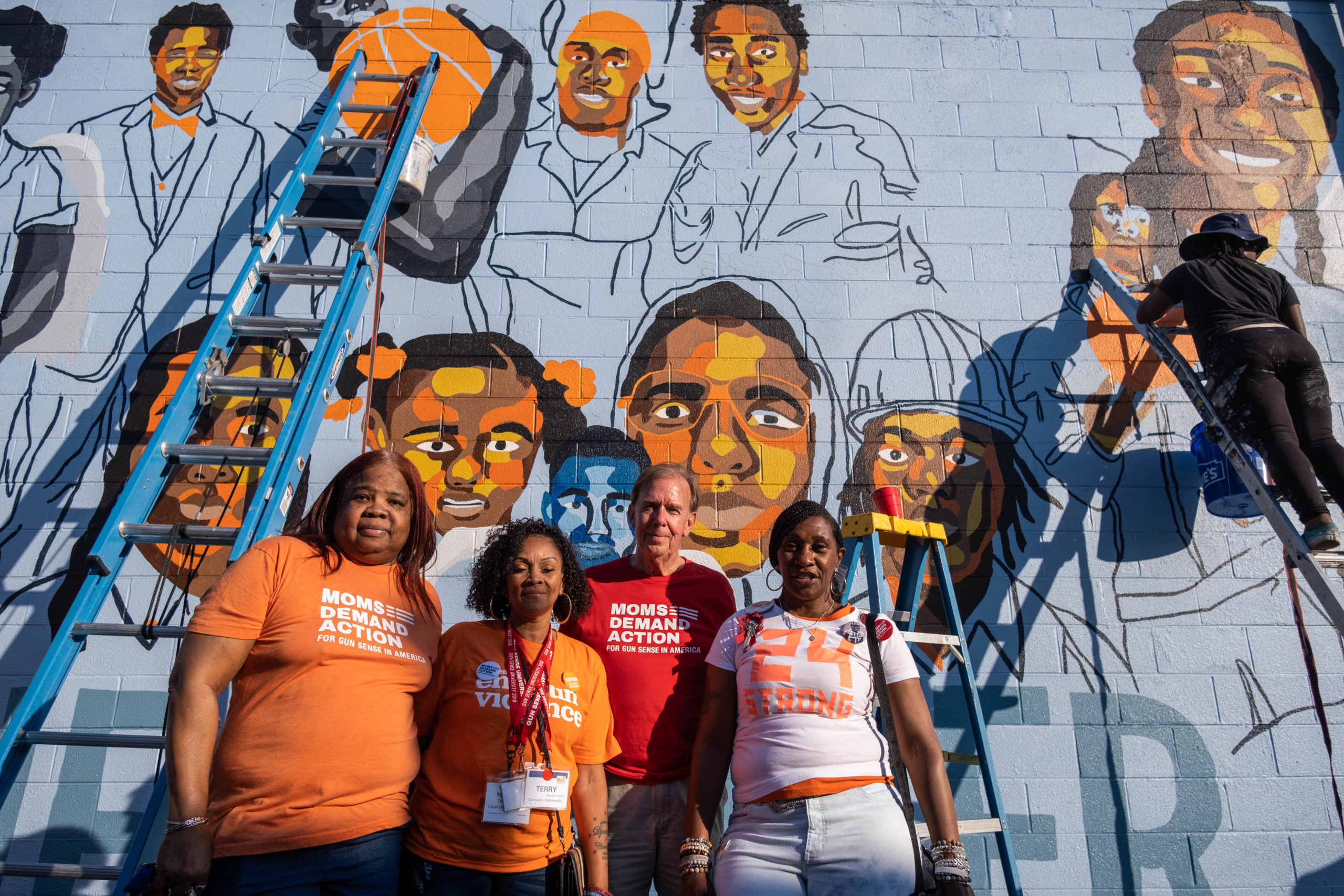 People in Wear Orange and Moms Demand Action t-shirts pose in front of a mural