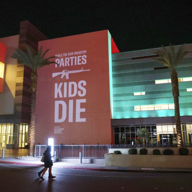 Photo of person walking by SHOT Show wall installation: While the gun industry parties, kids die