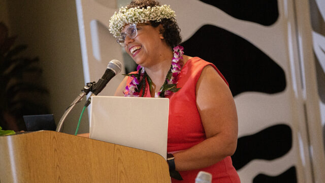 An image of Angelina Mercado, a woman with short, curly dark hair, wearing a white crown of baby's breath and a lei around her neck with magenta and white flowers. She wears a red dress and smiles as she speaks into a microphone at a podium.