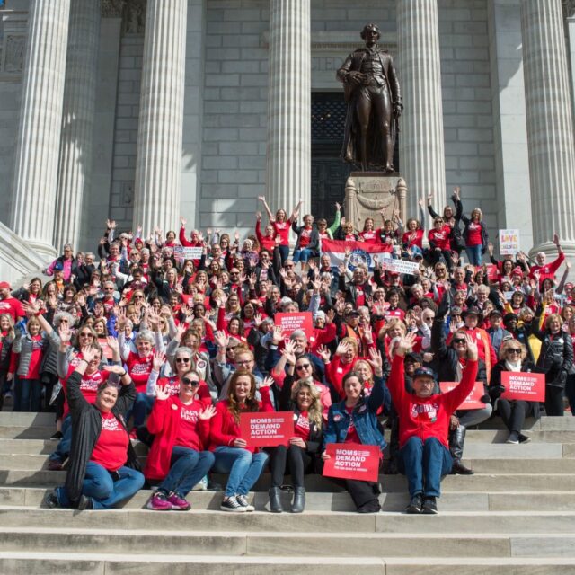A group of over 50 Moms Demand Action Volunteers cluster on the front steps of a government building, wearing red t-shirts, jeans, and holding Moms Demand Action red signs.