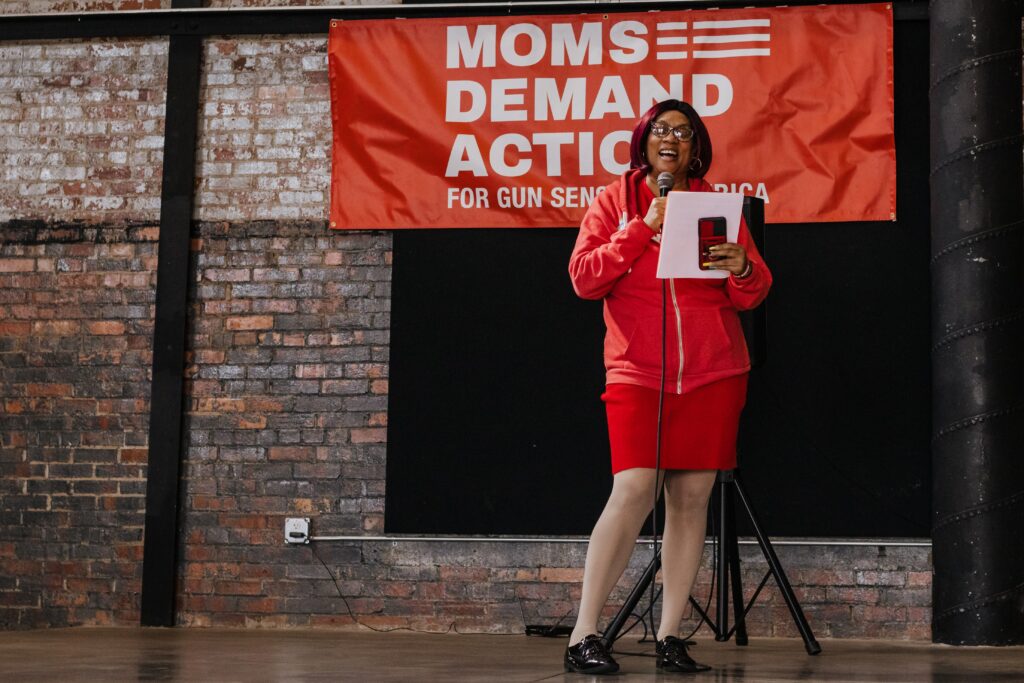 Leslie Anne Washington, a Black woman wearing a red zip-up sweatshirt, a red skirt, white tights, and black shoes, stands in front of a red "Moms Demand Action for Gun Sense in America" banner and reads from a white piece of paper. She holds a microphone in her right hand.
