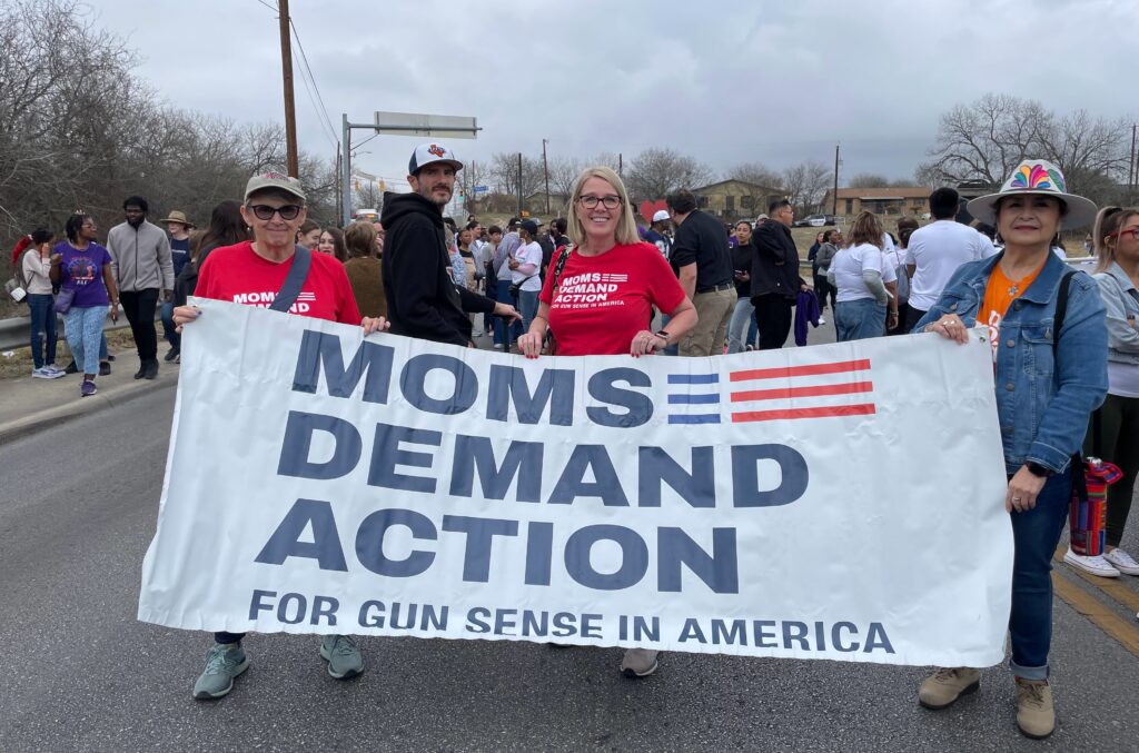 Moms Demand Action volunteer Jeni Olson, a white woman with a blond bob, wears a red shirt and stands behind a white banner that reads "Moms Demand Action for Gun Sense in America" in navy blue letters.