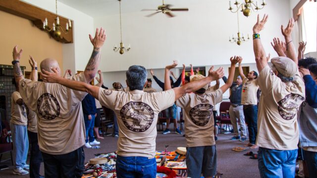 A group of retiro (retreat) participants gather in a circle. They wear denim pants and taupe t-shirts; their arms are stretched up toward the ceiling.
