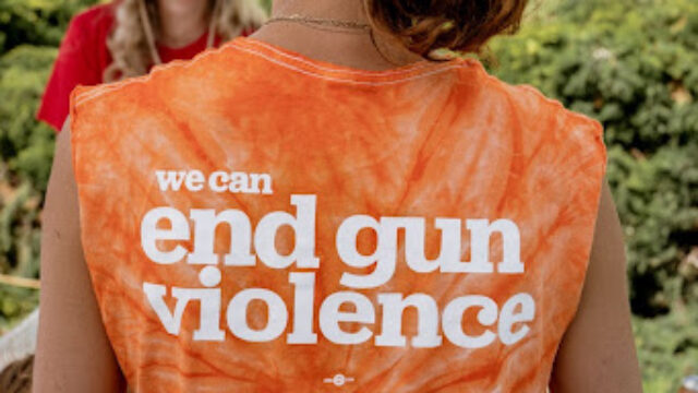 A volunteer is seen from the back. She wears a white mesh baseball cap and has her hair in a short ponytail that brushes the back of her neck. Her shirt is orange tie-dye and reads