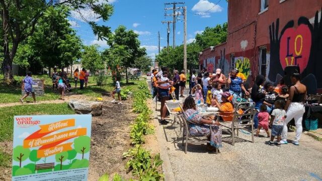 Sandtown community members gather at a pocket park in Baltimore, MD. A Wear Orange sign is visible in the foreground.