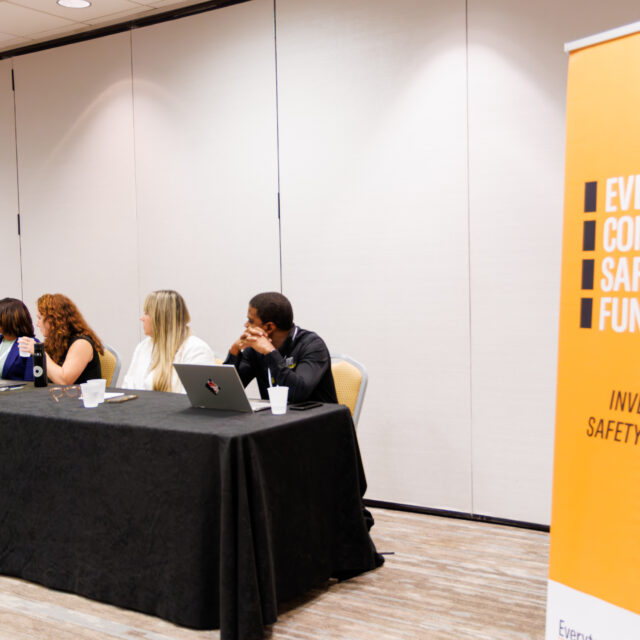 Five panelists sit at a table with a black tablecloth; to their right is an orange placard that reads
