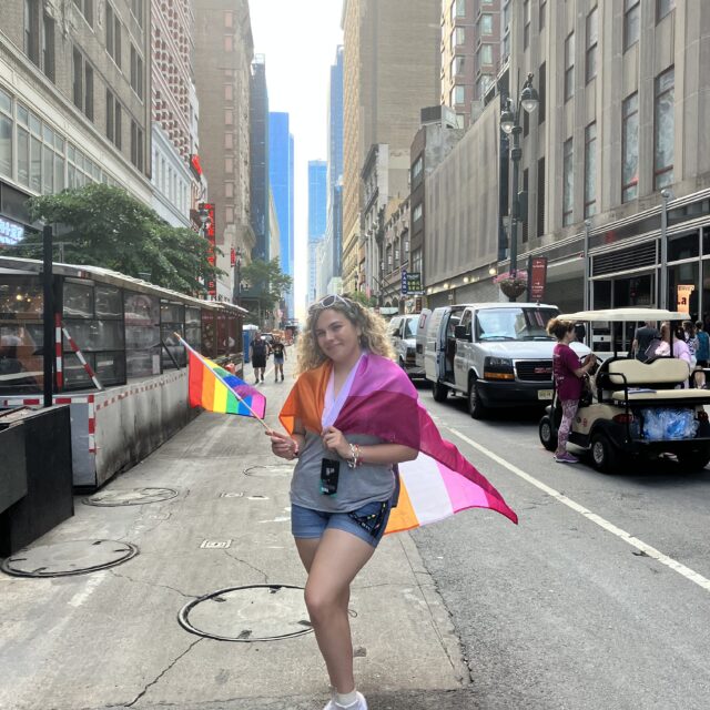 A person with light skin and shoulder-length curly blond hair poses for a photo int he middle of a street. Grey tall buildings are pictured on either side of them and recede into the distance. They have white sunglasses in their hair and hold a rainbow flag in their right hand. They have the lesbian pride flag wrapped around their shoulders. They wear a grey t-shirt, blue denim shorts, and white shoes with a rainbow on the side.