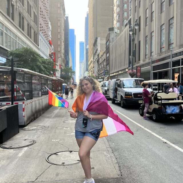 A person with light skin and shoulder-length curly blond hair poses for a photo int he middle of a street. Grey tall buildings are pictured on either side of them and recede into the distance. They have white sunglasses in their hair and hold a rainbow flag in their right hand. They have the lesbian pride flag wrapped around their shoulders. They wear a grey t-shirt, blue denim shorts, and white shoes with a rainbow on the side.