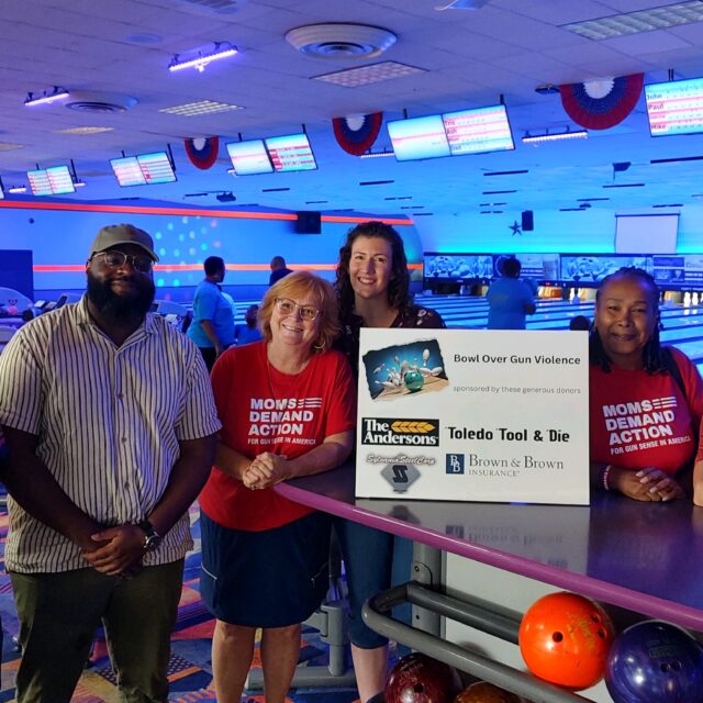 Six people pose for a photo inside of a bowling alley. Two are wearing Moms Demand Action shirts. They all are gathered around a white sign that reads