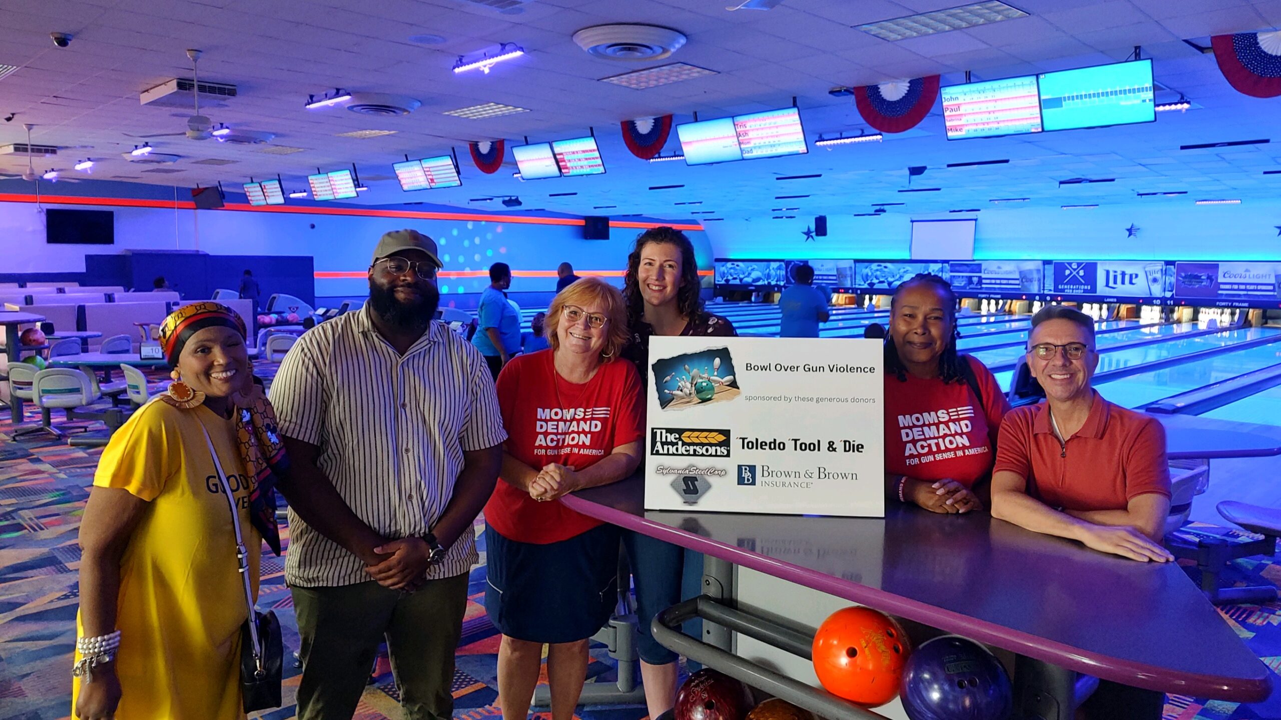 Six people pose for a photo inside of a bowling alley. Two are wearing Moms Demand Action shirts. They all are gathered around a white sign that reads "Bowl Over Gun Violence."