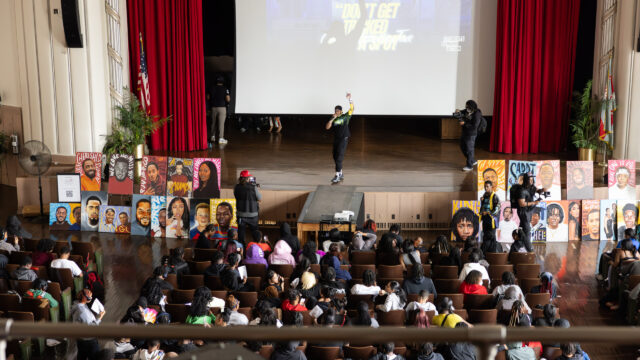 Manny, a Black man in his mid-20s, stands in the center of a brown stage in front of a projected slide that reads