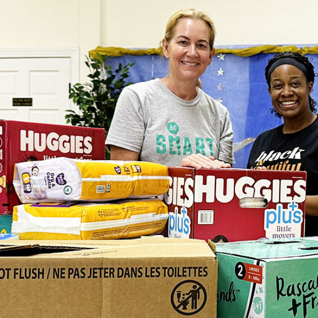 Shannon, a white woman with blond hair wearing a grey Be SMART t-shirt, poses with Crystal, a Black woman wearing a black t-shirt and a black headband. In the foreground of the image are boxes of diapers they have collected.