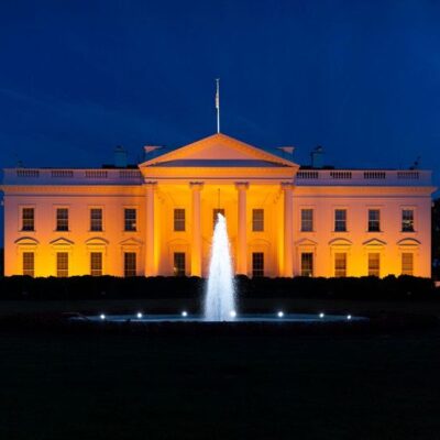 A front-facing view of the White House taken at night. A fountain is bubbling in the center of the shot, bisecting the White House. The White House is lit up the color orange for gun violence awareness. The sky behind it is navy blue.