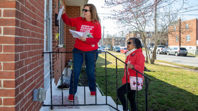 Two Moms Demand Action volunteers, wearing red long-sleeve shirts, are door-knocking. One stands at the top of a short staircase, left hand raised to knock on the front door of a brick building while her right hand holds a stack of elections pamphlet materials. The other stands at the bottom of the stairs, with her right leg one stair higher than her left. Her hands are in her pockets.