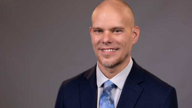 Jeremy Flowers, a white man wearing a navy blue blazer, a white collared shirt, and a gradient blue tie, smiles at the camera. He is pictured in front of a grey professional photo background.