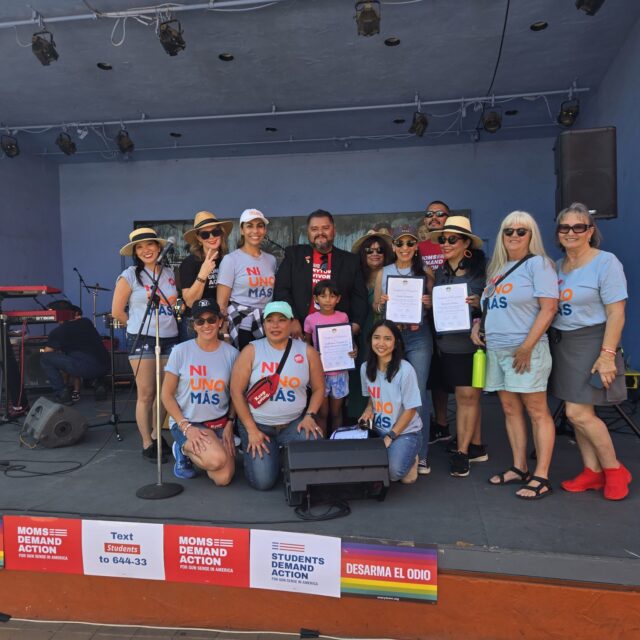 A group of 13 people stand on a stage and pose for a photo. Almost everyone is wearing blue t-shirts or grey tank tops that say