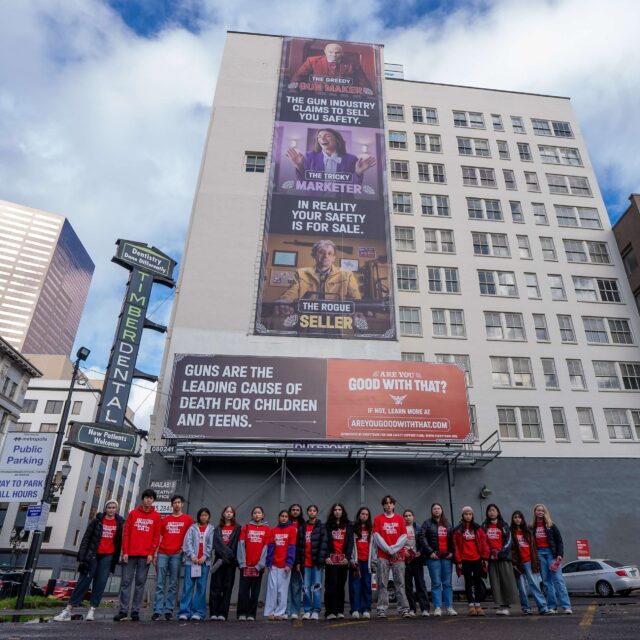 18 Students Demand Action volunteers pose for a low-angle photo. All are wearing red Students Demand Action t-shirts, pants, and jackets or long-sleeve layers. Behind the students, an