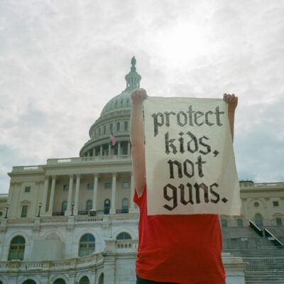 A low-angle photo of a person standing in front of the steps of the Capitol Building in Washington, D.C. The person is wearing a red shirt and is holding up a rectangular sign in front of their face that reads 