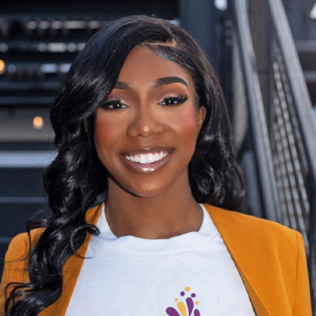 Aaliyah Strong, the founder and executive director of Tyme to Thrive Beyond Grief, poses for a portrait photo. Her black hair is parted on the side and she is wearing subtle glamorous makeup. She is facing the camera directly and is pulling her orange blazer slightly away from her white t-shirt so the full