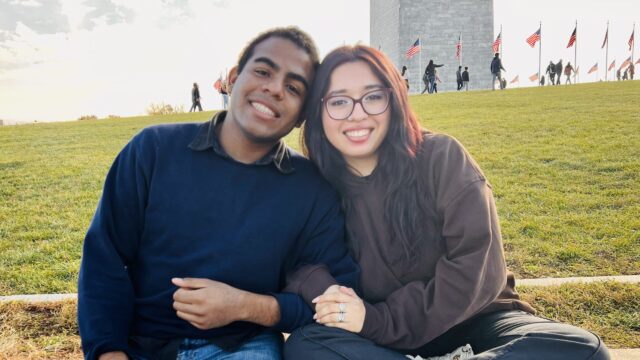 Kyle Clarke Echeverría and Andrea Joseline Rodríguez Ávila pose for a photo while sitting on a sidewalk in front of the Washington Monument. They are smiling and are linking arms.