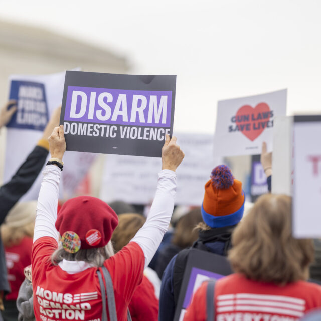 A view from the back of a crowd of Moms Demand Action volunteers protesting in front of the Supreme Court. One volunteer holds a sign that reads