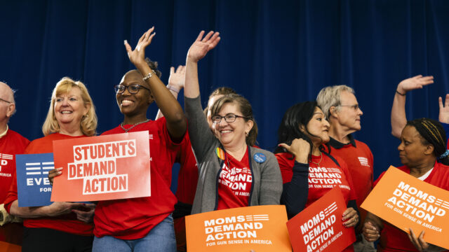 A group of people stand in front of a curtain at a rally. They are wearing red Moms Demand Action and Students Demand Action shirts and holding signs that say Everytown for Gun Safety, Students Demand Action, and Moms Demand Action for Gun Sense in America. CREDIT: Alyssa Schukar for Everytown
