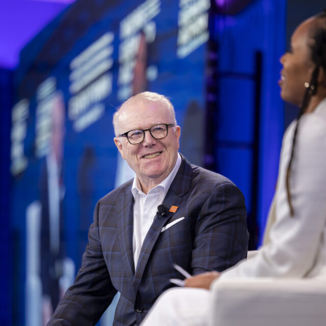 John Feinblatt, president of Everytown for Gun Safety Action Fund, wears a navy and black plaid suit, dark glasses, and a white collared shirt while speaking on a stage with Angela Ferrell-Zabala, executive director of Moms Demand Action.