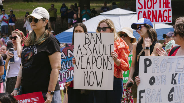 Gun safety advocates gather at a protest for stronger gun laws. One person holds a white sign with black text that reads 