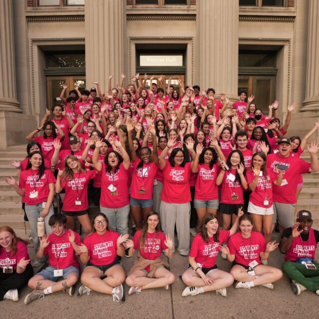A group of 100 Students Demand Action volunteers, all wearing red t-shirts with the Students Demand Action logo, gather for a photo on the Vincent Hall steps at the University of Minnesota during the Summer Organizing Institute. Students are raising their arms in the air and waving at the camera.