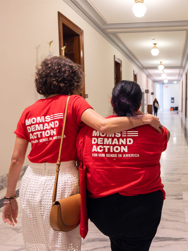 Two Moms Demand Action volunteers, wearing red t-shirts with the Moms Demand Action logo, are pictured from behind walking down a hallway during a Moms Demand Action Advocacy Day. One volunteer has her arm around the other.